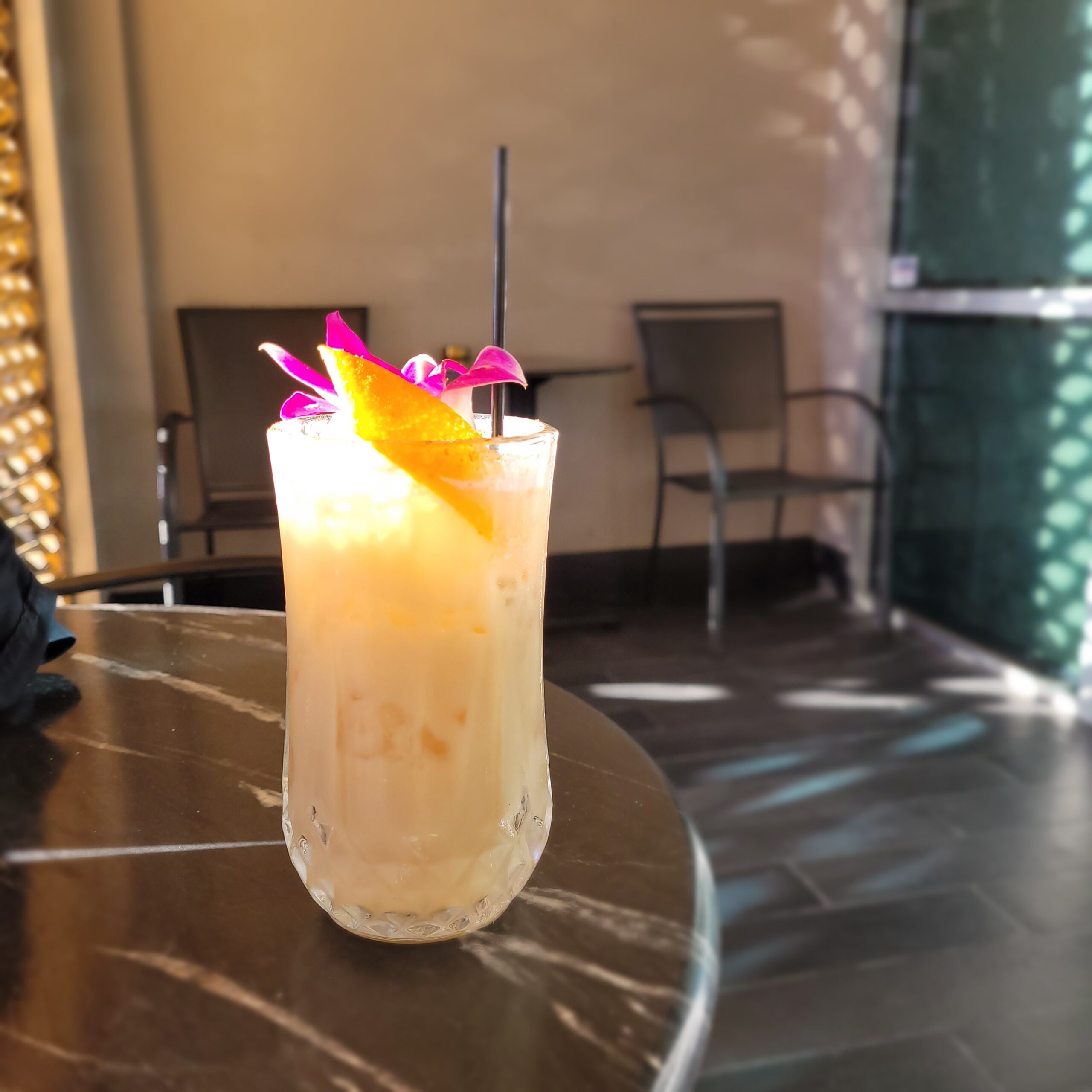 a tropical cocktail with a lemon duke, orchid garnish sits on a table with natural sun light on a table with empty chairs in the background