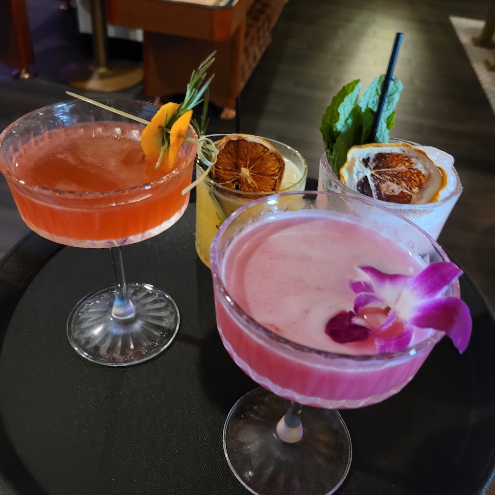 four vibrant cocktails on a cocktail tray on display with various garnishes from orchid flowers to mint, orange wedges, and rosemary in an indoor bar setting