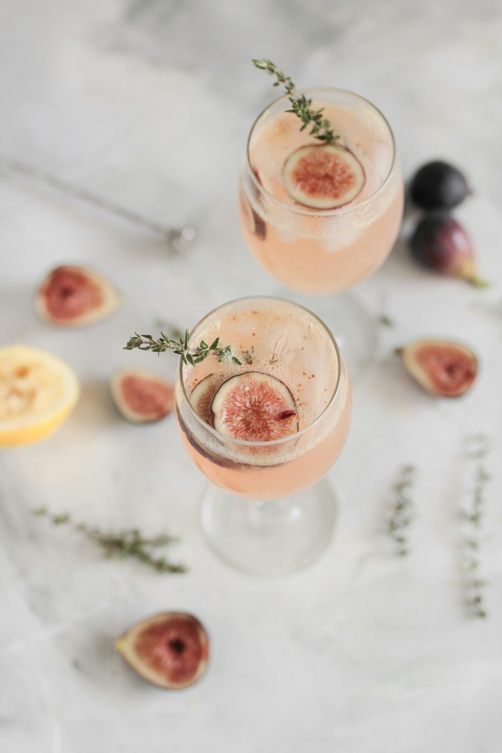 two fig cocktails with rosemary garnishes are displayed over a marble countertop with decorated figs and sliced lemons