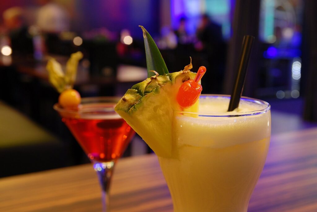 a tropical and cranberry cocktail are both displayed in an indoor bar setting that appears slightly upscale with neon lighting and curtains in the background