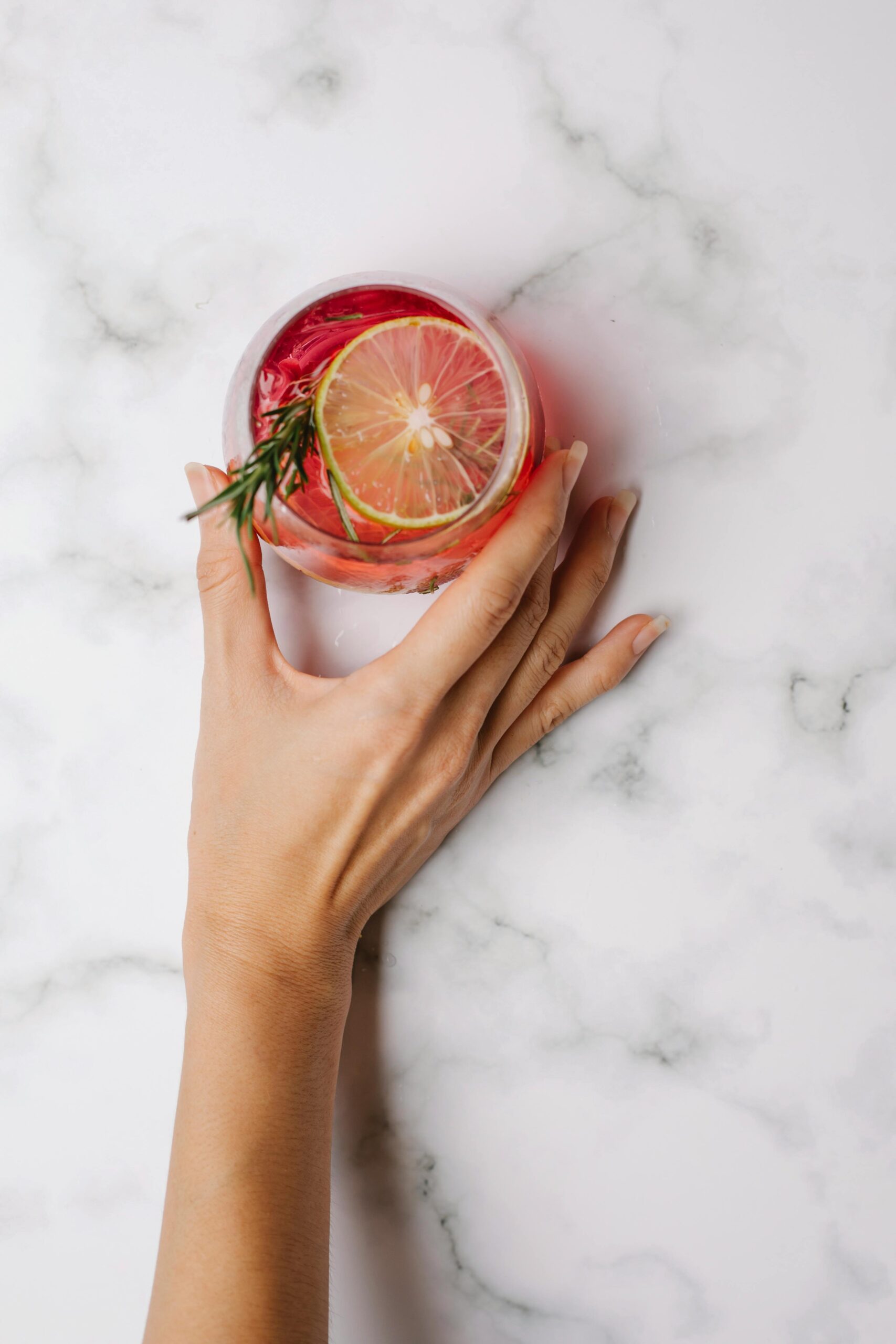 a woman's hand presents a cranberry cocktail from an aerial view over a marble countertop with lime and rosemary garnish