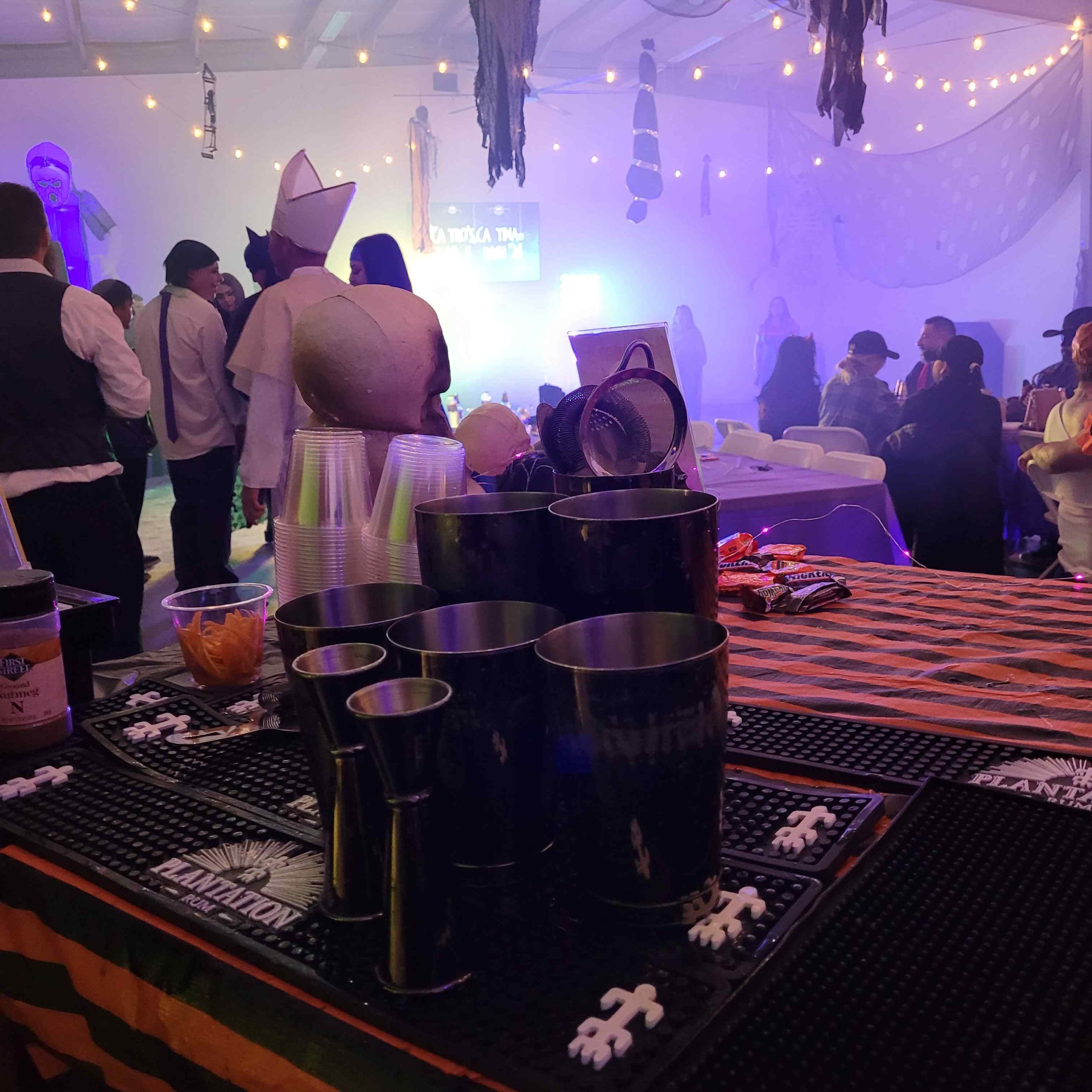 the bar setup showing cocktail shakers, cups, candy, and festive halloween decor before a group of costumed people socializing at a party